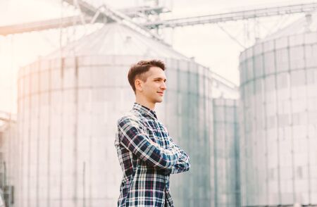 Portrait of confident business man owner of granary facilities keeping arms crossed while standing against silos buildings. Young successful agriculture industry business entrepreneur, grain storageの写真素材