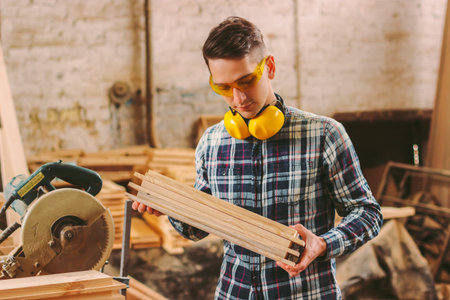 Young male carpenter in protective glasses and headphones holding stack of wooden planks in hands...