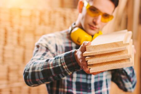 Young professional male joiner in protective goggles, headphones holding wooden planks in hands while working at sawmill factory. Confident skilled carpenter at wood production workshop or warehouseの写真素材