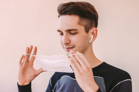 Portrait of happy young sports man listening music in wireless earphones and wearing medical face...