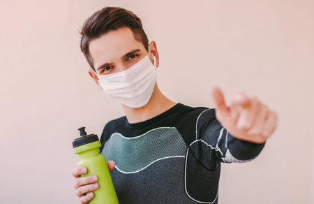 Confident sports man in medical face mask pointing with index finger and listening music. Happy young man fitness instructor in protective mask. Motivation for home training, COVID-19 quarantineの写真素材