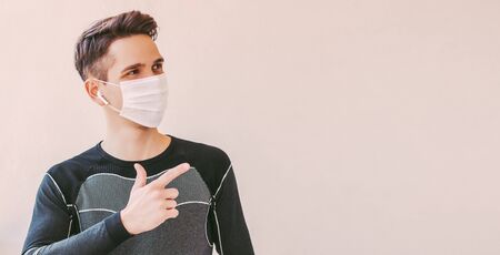 Young confident sports man in medical face mask pointing with finger to background with copy space. Happy fitness trainer in protective mask showing copy space. COVID-19 coronavirus home quarantineの写真素材
