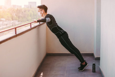 Young athletic sports man in protective face mask doing push ups exercises at home. Sporty confident...