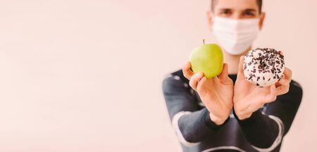 Young fitness man in medical face mask struggling between healthy and junk food. Happy sports man in protective mask hold sweet donut and fresh apple in hands. COVID-19 quarantine dieting, copy spaceの写真素材
