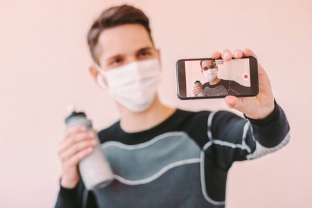 Young man fitness instructor in medical protective mask holding mobile phone in hand while streaming online video. Happy sports man recording video tutorial for training at home. COVID-19 quarantineの写真素材