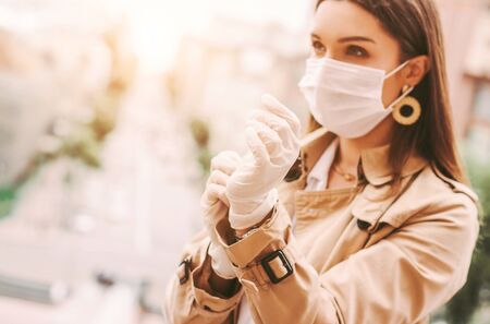 Beautiful young stylish woman in protective face mask put on medical latex gloves outdoors. Trendy elegant hipster girl in medical face mask wear protective gloves on city street. COVID-19 protectionの写真素材