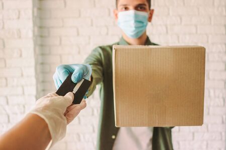Happy hipster delivery man in medical face mask hold cardboard box in hand, checkout customer. POV customer in rubber protective gloves receive shipment box from courier. COVID-19 quarantine deliveryの写真素材