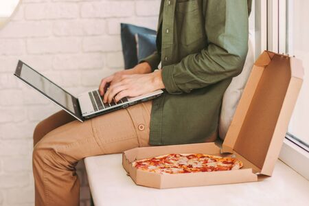 Closeup stylish hipster man typing laptop keyboard, eat delicious pizza, sit on windowsill. Busy businessman freelancer work remotely from home office and eat takeaway italian fast food. Lunch breakの写真素材