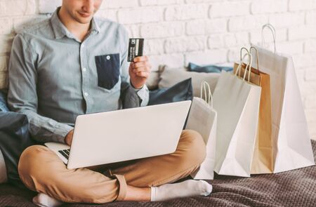 Closeup young happy man sit on couch, using laptop for safe online shopping from home. Stylish male hipster shopper hold credit card in hand. Payments and purchases at internet store. Man shopaholicの写真素材