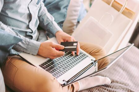 Closeup man shopper hold credit card in hand, typing on laptop keyboard, shopping online from home. Stylish hipster man sit on couch, using computer for internet payment, purchase. E-commerce storeの写真素材