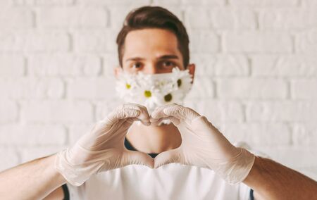 Portrait happy male doctor in protective gloves and flower medical face mask gesturing love sign with fingers. Young man in summer medical face mask showing heart shape symbol. Good, charity COVID-19の写真素材