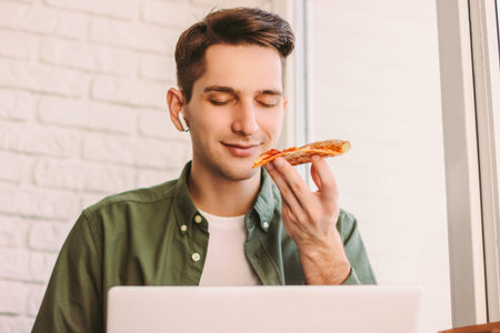 Hungry hipster man freelancer smell, hold slice of tasty italian pizza while study remotely on...