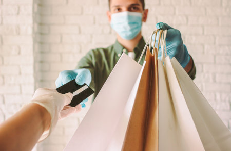 POV girl in protective gloves receiving shopping bags from delivery man in medical face mask. Male...