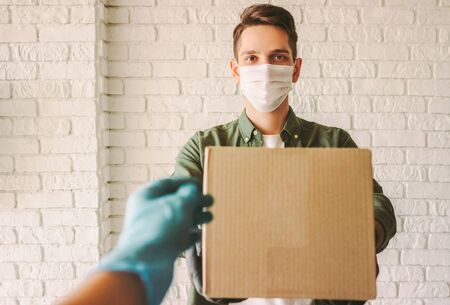 POV customer in rubber protective gloves receiving shipment box from delivery service employee. Happy hipster man courier in medical face mask give cardboard box to customer. Safe quarantine deliveryの写真素材