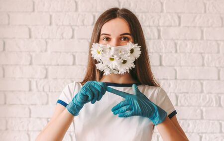 Portrait young girl doctor in medical face mask with flowers remove protective gloves from hands. Happy beautiful woman in protective mask celebrate end of coronavirus COVID-19 fight, take off glovesの写真素材