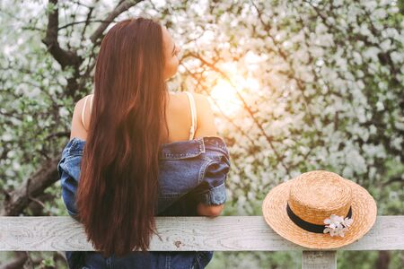 Back view of beautiful young brunette woman in denim jacket posing with wooden fence on countryside garden. Happy attractive hippie girl with long hair in stylish dress on sunny blossom meadowの写真素材