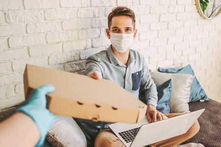 POV of delivery service employee in protective gloves giving cardboard pizza box to happy customer. Hipster man in medical face mask receive italian fast food from courier, work at home. Delivery foodの写真素材