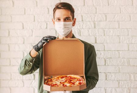 Hipster guy courier in medical face mask and gloves hold cardboard pizza box in hands. Happy man delivery service worker in protective mask carry open tasty pizza box. Italian fast food. Food deliveryの写真素材