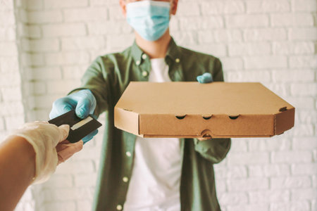 Hipster guy delivery service worker in medical face mask and gloves hold pizza box in hand, checkout...