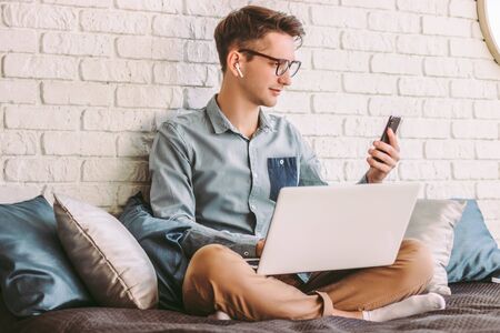 Young businessman in glasses hold, using mobile phone, working on laptop at home office. Stylish hipster man student text message on smart phone. Online communication, remote, distance job on couchの写真素材
