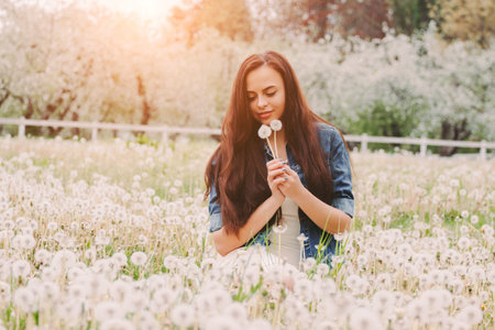 Portrait young beautiful hipster woman in denim jacket blow dandelions in hands while sit on green grass on garden. Happy carefree hippie girl relaxing on dandelion field. Spring flower pollen allergyの写真素材