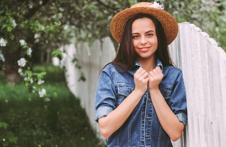 Portrait young happy hipster woman in stylish straw hat and denim jacket posing with wooden fence on green sunny meadow. Attractive cheerful hippie girl on countryside garden with blossom treesの写真素材
