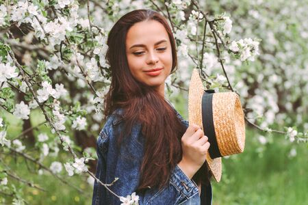 Portrait young gentle hipster girl in stylish denim jacket posing on countryside garden with blossom floral trees. Beautiful hippie woman relax and smile on green meadow. Enjoy spring nature outdoorsの写真素材