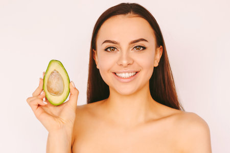 Portrait cheerful young girl with perfect clean smooth skin posing with avocado slice in hand, smile...
