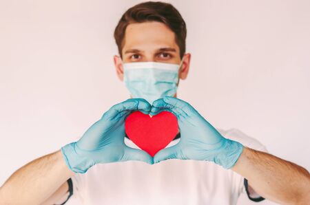 Portrait young man surgeon in medical face mask, protective gloves hold red heart in hands isolated on white background. Happy male doctor nurse gesture heart love sign. Health care, charity, donationの写真素材