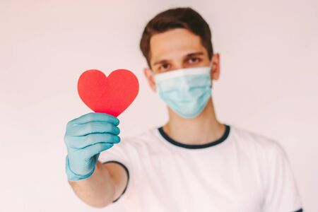 Happy young male doctor nurse in protective glove and medical face mask hold red heart symbol in hand isolated on white background. Man surgeon with love heart sign. Charity, donation, health careの写真素材
