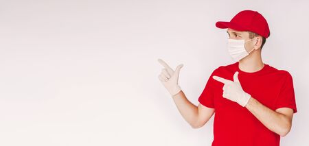 Banner happy delivery man in medical face mask and protective gloves point index finger to empty copy space on white background. Young delivery service employee in protective mask and gloves isolatedの写真素材