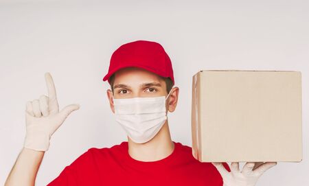 Portrait happy man delivery service employee in protective face mask, medical gloves, uniform point index finger on white background copy space. Young delivery guy carry cardboard box. Man courierの写真素材
