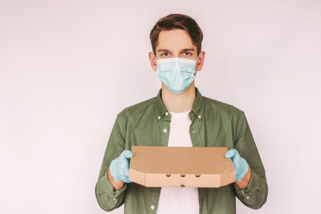 Portrait young delivery man in medical face mask and protective gloves hold cardboard pizza box in...