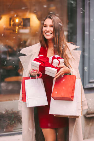 Cheerful beautiful girl with shopping bags, cane candy, gift boxes in hands smile outdoors. Happy young attractive woman in shopping mall. Christmas shopping, New Year Eve, winter holidays preparationの写真素材