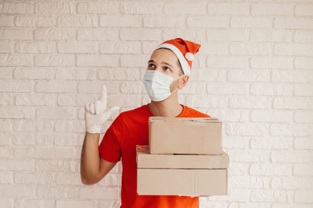 Young man courier in protective face mask, medical gloves, santa hat carry delivery boxes in hands. Happy delivery service employee in medical face mask hold cardboard packages. Christmas, New Yearの写真素材