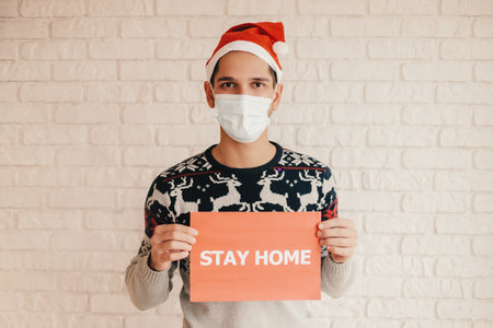 Male nurse in Santa hat, medical gloves, protective face mask show stay home message. Happy man doctor in face mask hold social awareness banner stay home in hands. Christmas self isolation campaignの写真素材