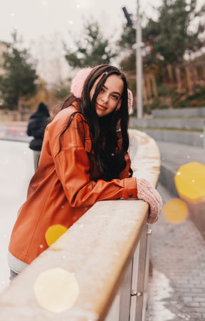 Portrait pretty young happy woman in stylish jacket, pink earmuffs skate on outdoor Christmas ice rink. Beautiful cheerful girl have fun and smile on ice skating rink. Winter holidays season activityの写真素材