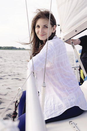 Happy smiling young woman wearing a white shirt and blue trousers, sitting. Sailboat trip.の写真素材