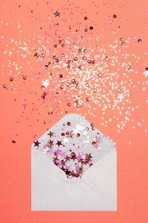 Sparkling confetti pouring out of white envelope on coral background. View from above. Flat lay. Holiday, congratulation or good news concept.の写真素材
