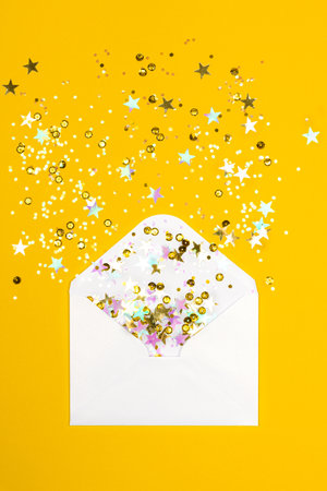 Sparkling confetti pouring out of white envelope on yellow background. View from above. Flat lay. Holiday, congratulation or good news concept.の写真素材