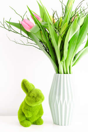 Bouquet of multicolored unopened tulips in light green ceramic vase and Easter green grass bunny on white wall background. Holiday decoration concept.の写真素材