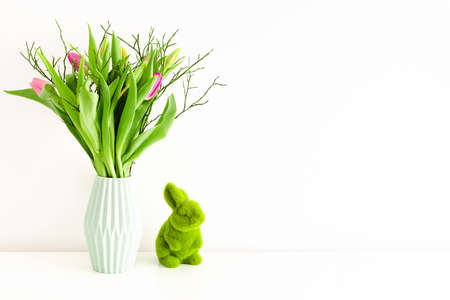 Bouquet of multicolored unopened tulips in light green ceramic vase and Easter green grass bunny on white wall background. Holiday decoration concept. Place for text.の写真素材