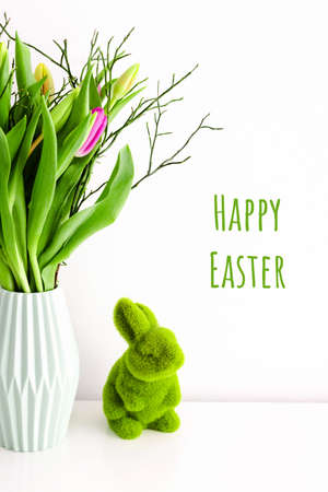 Greeting card with bouquet of multicolored unopened tulips in light green ceramic vase and Easter green grass bunny on white wall background. Holiday decoration concept.の写真素材