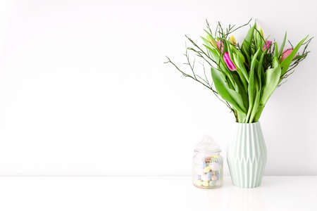 Bouquet of multicolored unopened tulips in light green ceramic vase and Easter candy jar filled with eggs on white wall background. Home interior decoration concept. Place for text.の写真素材