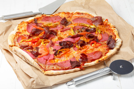 Pizza with ham, salami and chili pepper, on wooden table with lifter and cutter knifeの写真素材