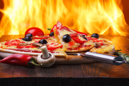 Pizza on wooden table with one slice on a lifter, real oven fire on backgroudの写真素材