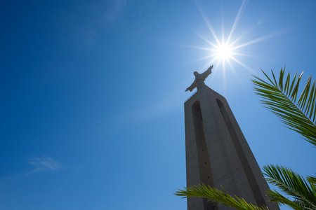 Huge statue of Christ the King through green palm leafs at clear summer sunny day in Lisbon Portugal with copy spaceのeditorial素材