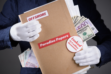 Man in suit with euro and dollar currency in pokets and in white gloves hold paper folder with Paradise Papers and Offshore label with documents inside, tax heaven leak conceptの写真素材