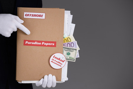 Man in suit and white gloves hold paper folder with Paradise Papers and Offshore label with documents and money inside, tax heaven documents leak conceptの写真素材