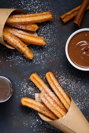 Churros with sugar powder chocolate sauce dip and cinnamon sticks on dark background flat lay top viewの写真素材